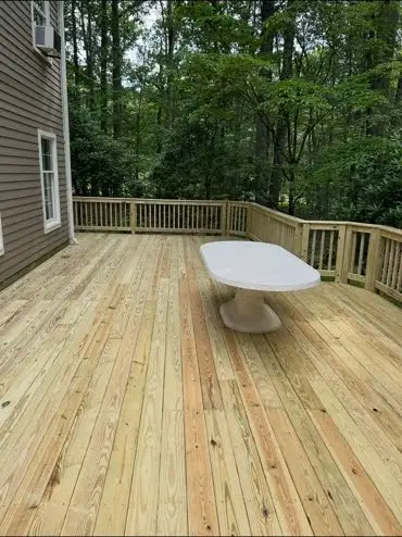 New unstained wood deck with railings attached to gray house, white oval table, woods behind.