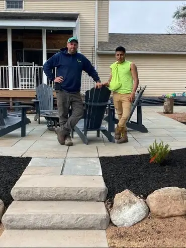 Randy in navy hoodie and teal cap and Ronaldo in green sleeveless shirt and tan pants on finished paver patio with Adirondack chairs, stone steps, mulch beds, house behind.