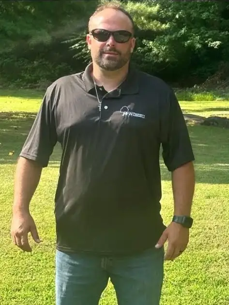 Randy, owner of RFW Property Management, stands on a mowed lawn wearing a black polo shirt, jeans and sunglasses, with trees behind him.